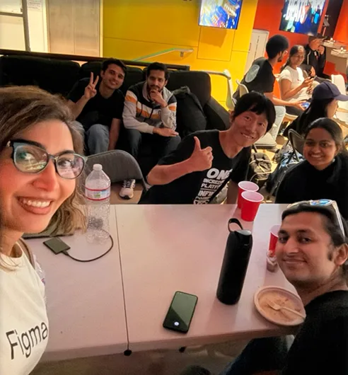 Group selfie of a hackathon team smiling around a table with laptops and snacks.