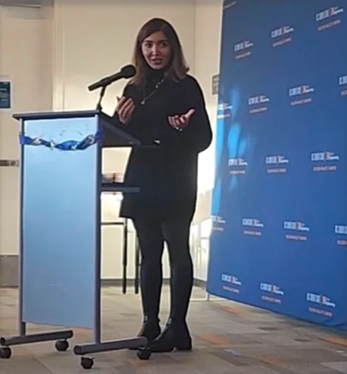 A woman speaking at a podium in front of a blue event backdrop.
