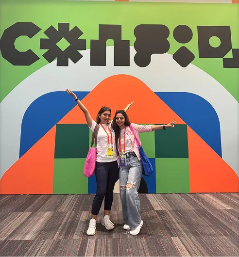 Two friends posing with arms outstretched in front of a colourful Config sign.